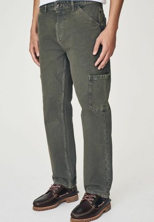Cargo pants in olive green, featuring a relaxed fit, side pockets, and a slightly distressed texture. Paired with lace-up dark shoes.