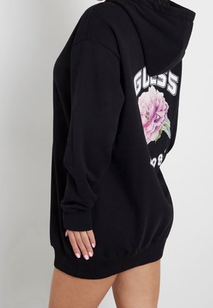 Person wearing oversized black hoodie with pink flower and "GUESS 1981" logo on the back, standing against a plain light background.