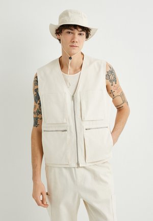 Beige, vetoketjullinen liivi, jossa on kaksi suurta etutaskua ja kaksi pienempää vetoketjutaskua, käytetty verkkopaidan päällä. Yhteensopiva beige buckethattu täydentää asun.
