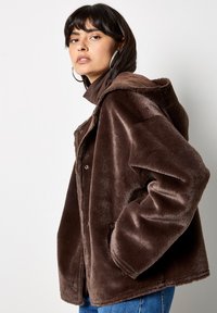 Giacca in pelliccia sintetica marrone con cappuccio, chiusure a bottoni a scatto e due tasche laterali. Presenta una texture morbida e una vestibilità oversize.