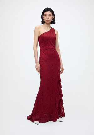 Femme portant une robe longue rouge en dentelle sans manches avec une épaule apparente et un détail volanté, debout devant un fond blanc uni.