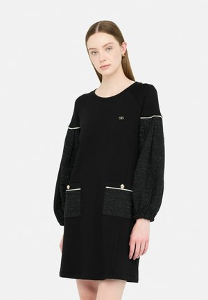Robe noire avec des manches bouffantes pailletées noires, présentant des accents en métal doré et deux poches avant avec des détails de boutons.