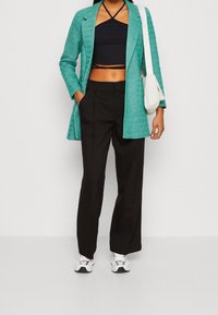 Türkiser strukturierter Blazer, schwarzes Crop Top, hochgeschnittene schwarze Hose und weiße Sneakers. Eine kleine weiße Umhängetasche rundet den Look ab.