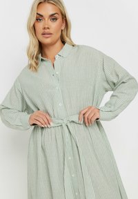 Grönvit randig skjortklänning med långa ärmar, krage, knäppning framtill och ett knytbart tygbälte i midjan.