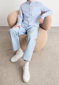 Hellblaues Hemd mit Knopfleiste und gesticktem Logo, kombiniert mit hellen Jeans und weißen Sneakers. Der Sitz ist beige, strukturiert und rund.