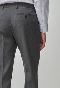 Pantalons gris sur mesure avec un tissu texturé, comportant une poche arrière avec un bouton et un design ajusté, présentés de profil.