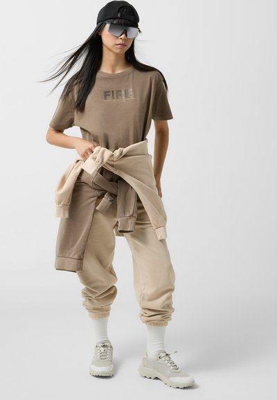 Beige cotton t-shirt with "FIRE" graphic, paired with beige joggers. Accessories include a black cap and oversized sunglasses. White socks with sneakers.