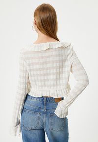 Weißer, kurzer Pullover mit strukturiertem Strickdesign, Rüschensaum am Halsausschnitt und langen Ärmeln. Kombiniert mit blauen Jeans aus Denim mit Gesäßtaschen.