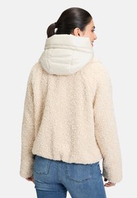 Veste en sherpa crème, douce et duveteuse, avec une capuche matelassée beige clair, affichant une coupe décontractée et des poignets côtelés. Vue arrière présentée.