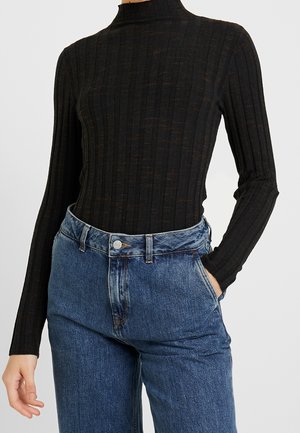 Personne portant un col roulé noir côtelé à manches longues rentré dans un jean bleu taille haute, une main dans la poche.