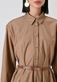 Bruine jurk met kraag en knoopsluiting, getailleerde taille en een bruine leren riem met een ronde metalen accent. Gladde, gestructureerde stof.