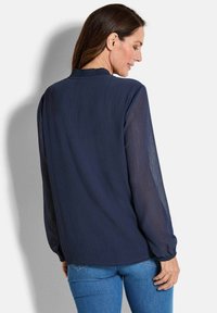 Marineblaue Bluse mit strukturiertem Finish, langen transparenten Ärmeln, gerafftem Ausschnitt und lockerer Passform, kombiniert mit hellblauen Jeans.