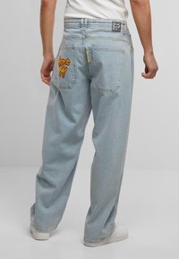 Hellblaue Denim-Jeans mit lockerer Passform. Sie haben eine rote "Home Boy"-Stickerei auf der Gesäßtasche und ein Etikett in der Nähe des Bunds.