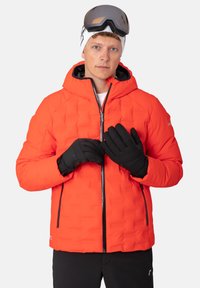 Rote isolierte Jacke mit einem strukturierten Design, Reißverschlusstaschen und Kapuze. Getragen mit schwarzen Handschuhen und einer Schutzbrille, vor einem schlichten Hintergrund.