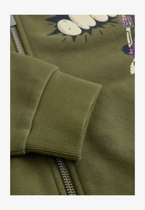 Olivno zelena jopa z zadrgo, s pritiski na rokavih, mehko teksturo in risanim motivom. Kovinska zadrga doda funkcionalnost.