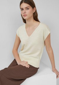 Jasny beżowy, krótki rękaw, dzianinowy top z dekoltem w kształcie litery V. Miękka faktura, żeberkowy dół. Połączony z brązowymi spodniami o fakturze, siedzący na białym piedestale.