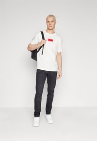 Vit t-shirt med röd grafik, i kombination med mörka jeans och vita sneakers. Modellen bär en svart väska över ena axeln, stående mot en enfärgad bakgrund.