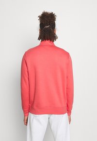 Korallfärgad sweatshirt med hög krage, ribbstickade muddar och fåll. Mjuk textur, avslappnad design. Kombinerad med ljusa byxor.