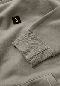 Grijs sweatshirt met ribgebreide manchetten, voorzien van een zwart vierkant label met een witte "J." Geweldige textuur en een ontspannen ontwerp.