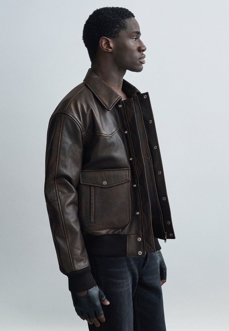 Καφέ δερμάτινο μπουφάν με γιακά, εμπρόσθιες τσέπες και ριμπ μανσέτες. Λεπτή υφή με ορατές ραφές. Φοριούνται μαύρα γάντια.