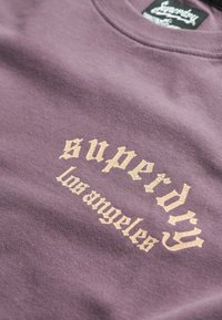 Paarse stof met beige gotische tekst met de woorden "superdry" en "los angeles" op een kledingstuk met ronde hals.