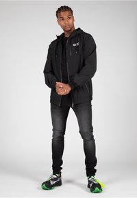 Giacca nera in nylon con zip e coulisse, indossata sopra una maglietta nera. Abbinata a jeans neri con ricami e scarpe da ginnastica nere e verdi.
