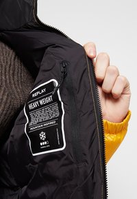 Schwarzes Innenfutter einer schweren Jacke mit gelbem Äußeren, mit einem Etikett auf der linken Seite und einem Reißverschluss auf der rechten Seite.