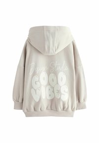 Beige huvtröja med dragsko, oversized passform och elastiska ärmmuddar. Baksidan är designad med vit text "Design Studio GOOD VIBES."