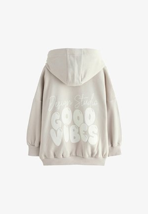 Smėlinė mikina su raišteliu, per dideliu siluetu ir elastingomis rankogaliais. Nugaroje yra balta užrašyta "Design Studio GOOD VIBES."