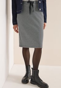Zwart-witte geruite potloodrok met een strik om de taille, gecombineerd met zwarte veterschoenen en doorschijnende zwarte panty’s.