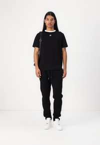 Svart t-shirt med vit rund halsringning och logotyp, tillsammans med svarta sweatpants och vita sneakers. Modellen bär också en svart axelväska.