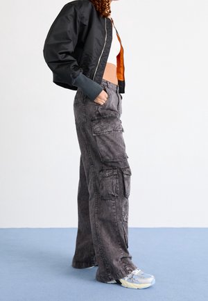 Persoon met een zwarte cropped bomberjack met oranje voering, een wit cropped topje, grijze cargobroek met meerdere zakken en witte sneakers.