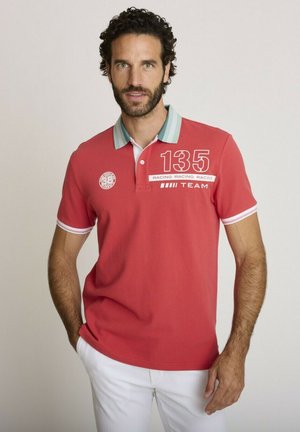 Homme aux cheveux bouclés foncés et à la barbe, portant un polo rouge avec les inscriptions « 135 » et « Racing Team », assorti d'un pantalon blanc, debout les mains dans les poches.