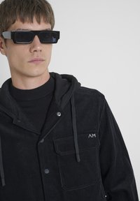 Chaqueta de pana negra con capucha, bolsillos delanteros, botones a presión y parche de logo plateado. Llevada sobre un suéter negro de cuello alto, combinada con gafas de sol negras.