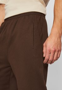 Pantaloni jogging din material moale, de culoare maro, cu talie elastică. Prezintă o buzunar lateral și o croială lejeră. Detalii cu cusături lângă buzunar.