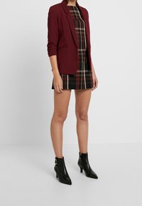 Burgundy blazer with rolled sleeves, paired with a black plaid mini dress. Model wears black pointed ankle boots with a side zipper.
