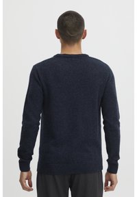 Pull en maille bleu marine, design col rond, poignets et ourlet côtelés, texture douce, porté avec un pantalon gris, modèle vu de dos.