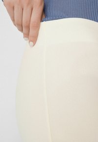 Pantalon de couleur crème en tissu texturé. La taille est montrée, légèrement tenue par une main, mettant en valeur un bord lisse.