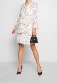 Vestido blanco en capas con lunares negros, mangas largas, escote en V y detalles de volantes. Modelo sosteniendo un pequeño bolso negro y usando tacones negros.