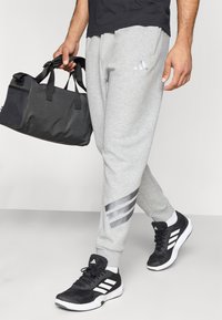 Grå sweatsbyxor med en åtsittande passform, prydda med ett silverfärgat tre-stripes mönster på vänster ben, i kombination med svarta träningsskor och en svart väska.