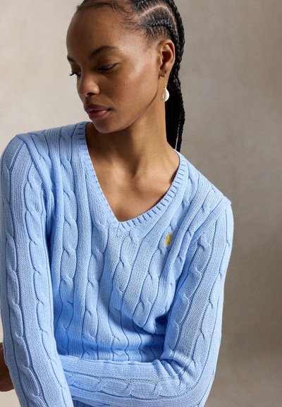 Žena s vrkočmi, oblečená v svetlomodrom svetríku s výstrihom do V a jedným visiacim náušnicou s mušľou, pozerajúca sa dole.