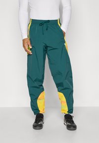 Pantaloni jogger turchesi con accenti gialli ai lati e toppe colorate sulle gambe. Realizzati in tessuto leggero e liscio con una vita elastica.