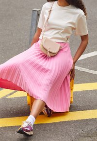 Gonna plissettata rosa, top beige e sneakers alla moda. Gli accessori includono una piccola borsa crossbody beige con un design testurizzato.