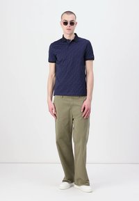 Marineblå polo med mønstrete tekstur, svart krage, sammen med løse olivengrønne bukser og hvite joggesko, iført runde solbriller.