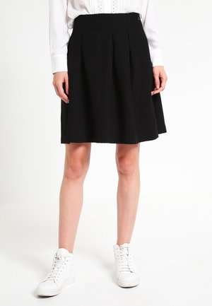 Femme portant une jupe plissée noire longueur genoux, un chemisier blanc à manches longues et des baskets blanches montantes, se tenant devant un fond uni.
