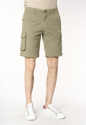 Uomo che indossa pantaloncini cargo beige con molte tasche e sneakers bianche con dettagli rossi, in piedi davanti a uno sfondo uniforme.
