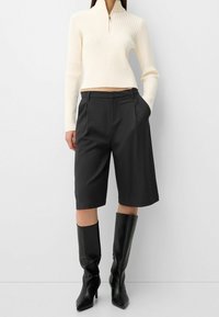 Nainen, jolla on päällä vaalean beige ribattu vetoketjullinen villatakki, tummat polvimittaiset räätälöidyt shortsit ja mustat korkea saappaat yksinkertaista taustaa vasten.