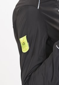 Sort løbejakke med en synlig gul LED-lysmodule og en tænd/sluk-knap på venstre ærme, lavet af letvægts, textured materiale.