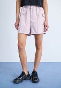 Pantaloni scurți roz deschis, realizați dintr-un material texturat; au un brâu elastic, buzunare laterale și o croială lejeră; purtați cu pantofi sport negri.