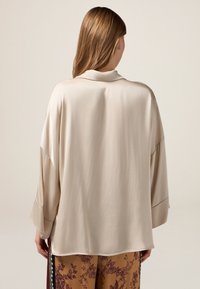 Blusa di seta beige dal taglio morbido, con maniche ampie e colletto ripiegato, caratterizzata da una texture liscia e un design minimalista.